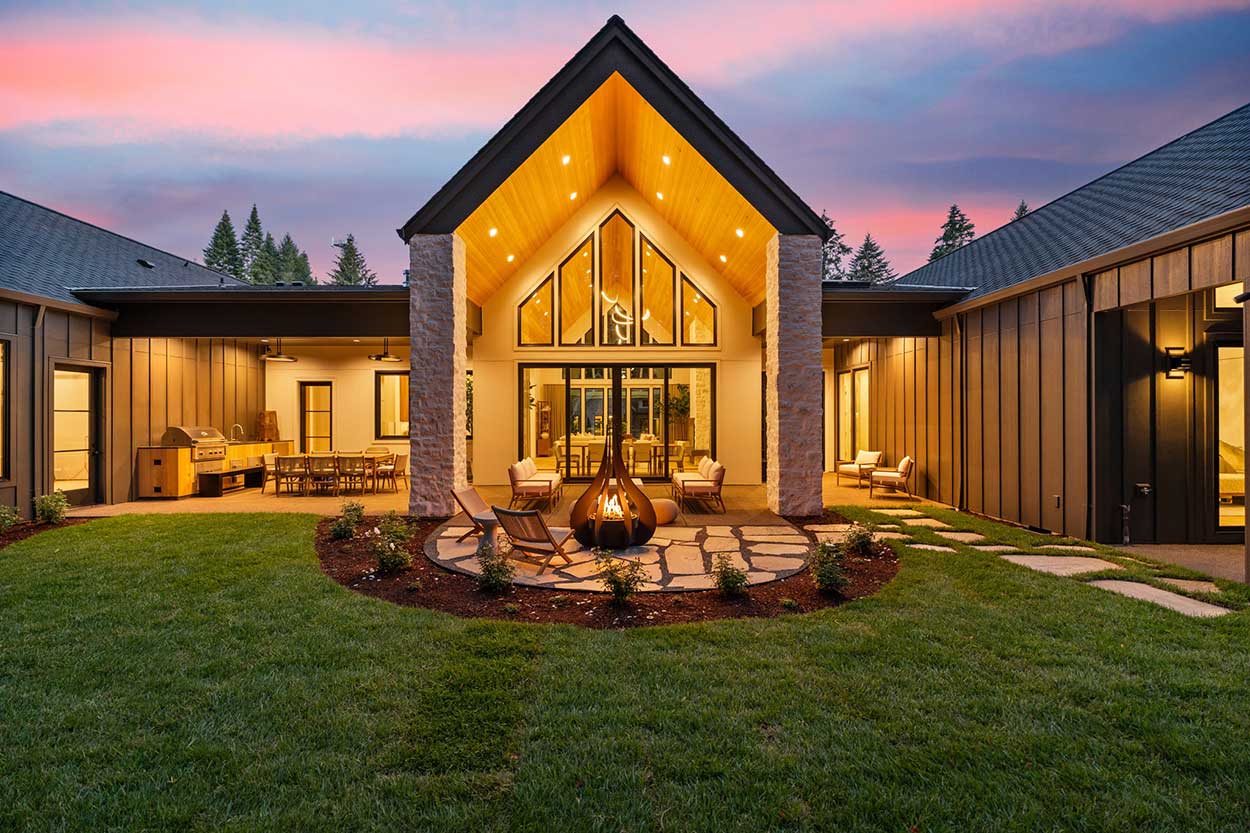 Rear courtyard at dusk with a lit fire pit, glowing gabled window wall, and outdoor kitchen and dining under covered patios.